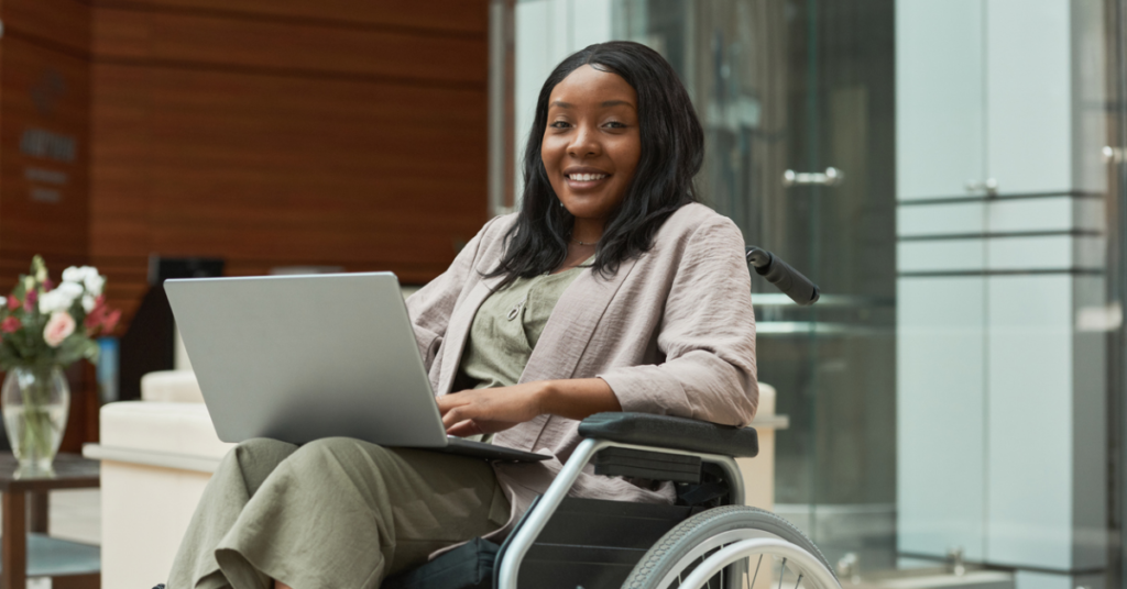 A photo of a woman in a wheelchair holding a laptop and smiling at the camera